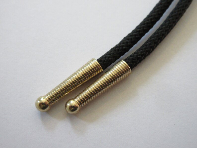 May include: Close-up of a black bolo tie with gold-toned metal tips. The tips have a ridged design.