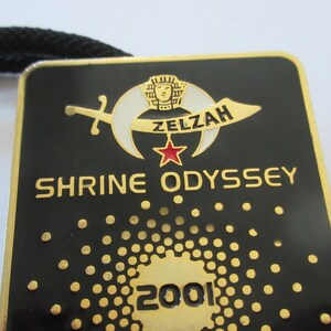 May include: A gold-colored metal pin with a black background. The pin features a crescent moon with a star and a sword, and the text "Zelzah Shrine Odyssey 2001".