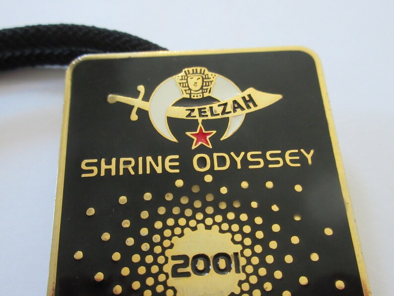 May include: A gold-colored metal pin with a black background. The pin features a crescent moon with a star and a sword, and the text "Zelzah Shrine Odyssey 2001".