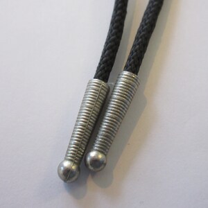 Puede incluir: Primer plano de dos cordones de corbata bolo negros con puntas de metal plateado. Las puntas tienen un dise&ntilde;o acanalado y un extremo redondeado.