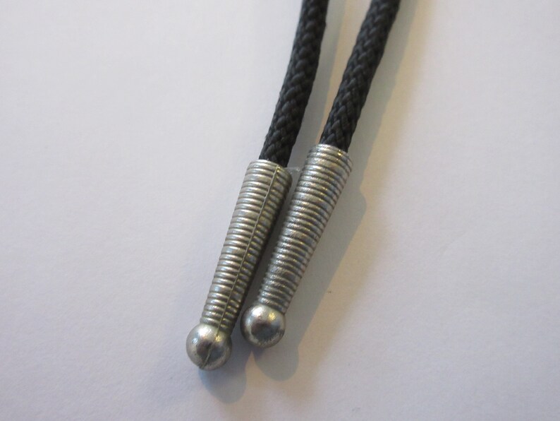 Puede incluir: Primer plano de dos cordones de corbata bolo negros con puntas de metal plateado. Las puntas tienen un dise&ntilde;o acanalado y un extremo redondeado.