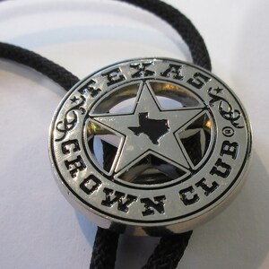 Puede incluir: Corbata bolo de plata con un colgante redondo negro y plateado que presenta una estrella de cinco puntas con el estado de Texas en su interior y las palabras "Texas Crown Club" alrededor de la estrella.
