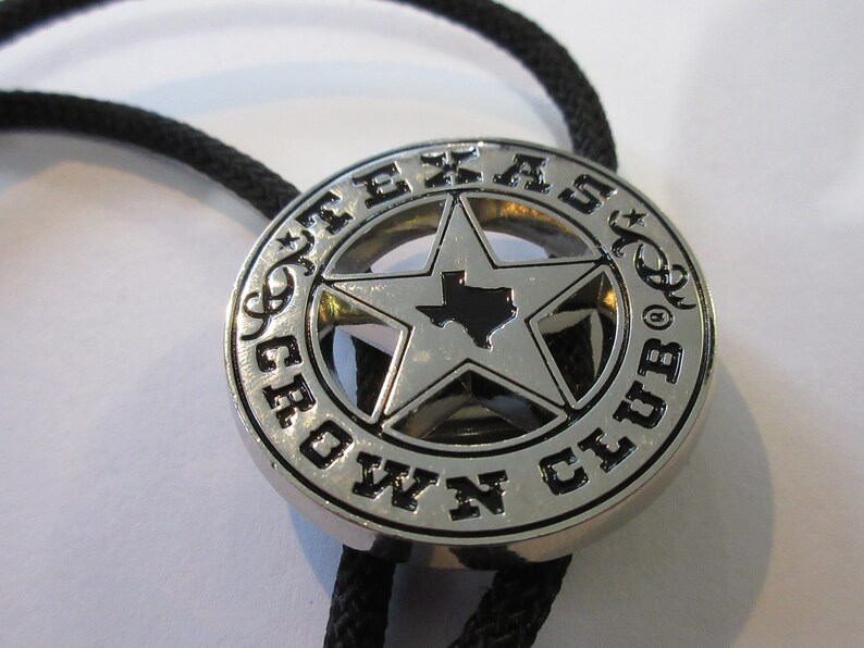 Puede incluir: Corbata bolo de plata con un colgante redondo negro y plateado que presenta una estrella de cinco puntas con el estado de Texas en su interior y las palabras "Texas Crown Club" alrededor de la estrella.