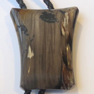 May include: A brown, black, and white patterned bolo tie with a rectangular pendant. The pendant is made of a natural material with a smooth, polished finish.