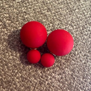 May include: A pair of red double-sided stud earrings. Each earring features a large, round, matte red ball paired with a smaller red ball. The earrings are set on silver posts.