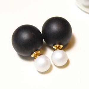 May include: A pair of black and white drop earrings. Each earring features a large, matte black sphere above a smaller, white, pearl-like sphere. The earrings have gold-colored posts. Additional black and white spheres are visible in a clear container.