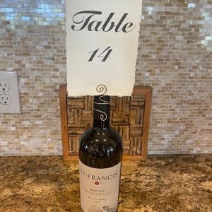 May include: A white card with black text that reads "Table 14" is attached to a wine bottle with a wire. The bottle is labeled "St. Francis Merlot".