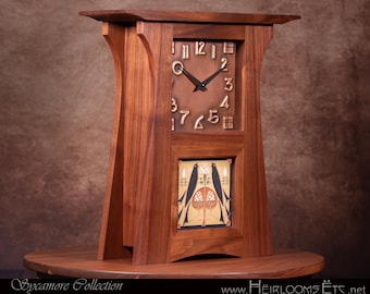 May include: A wooden mantel clock with a brown finish. The clock face has bronze-colored numerals and black hands. Below the clock face is a decorative panel with a bird design. The clock is on a round wooden table.