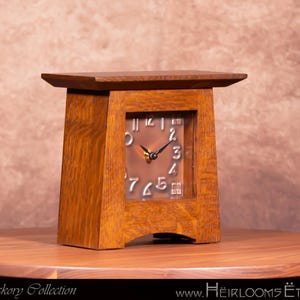 May include: A wooden table clock with a square face and a copper-colored clock face. The clock has a dark brown wooden frame and a light brown wooden top. The clock hands are black. The clock is sitting on a round wooden table.