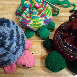 May include: Three handmade crochet turtle-shaped drawstring bags. One bag is blue with pink feet, another is multicolored with green feet, and the third is brown with green feet. The bags are on a wooden surface.