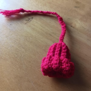 May include: A handmade, bright pink crocheted bell with a long, twisted cord and tassel. The bell shape has a ribbed texture. The tassel and cord are attached to the top of the bell. The item is on a wooden surface.