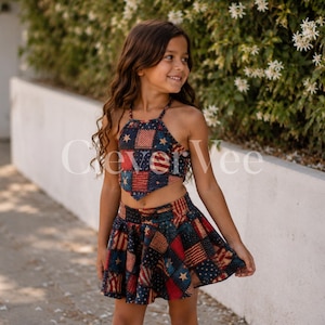 May include: A two-piece outfit featuring a halter top and a skirt. The top and skirt are patterned with a patchwork design in red, white, and blue, with stars and stripes. The outfit is designed for children.