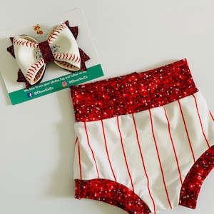 May include: Baseball-themed baby clothing set. Includes a white and red striped pair of shorts and a baseball-shaped bow. The bow is white with red stitching and a glittery red center. The shorts have a glittery red waistband.