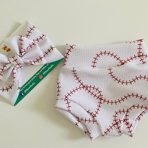 May include: White baby shorts and a matching headband, both featuring a baseball stitch pattern in red thread. The fabric has a textured appearance. The items are displayed on a white surface.