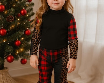 Op de afbeelding: Een kind draagt een outfit met een zwarte coltrui met rood geruite en luipaardmouwen. De bijpassende broek heeft een rood geruit en luipaardpatroon. Op de achtergrond een kerstboom met rode ballen.
