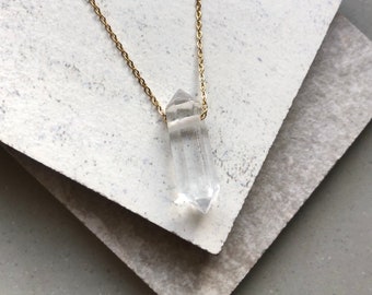 May include: A clear quartz crystal pendant necklace with a gold-colored chain. The crystal is faceted and elongated, suspended from a delicate chain. The necklace is displayed on a textured, layered surface, creating a minimalist aesthetic. The crystal measures approximately 2 inches in length.