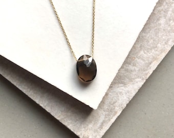 May include: A gold chain necklace featuring a faceted, oval-shaped smoky quartz pendant. The pendant is a translucent brown color and hangs from a delicate gold chain. The necklace is displayed on a white and beige textured surface.