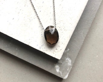 May include: A silver chain necklace featuring a faceted, oval-shaped smoky quartz pendant. The pendant is a translucent brown color and catches the light, creating a sparkling effect. The necklace is displayed on a textured white and gray surface.