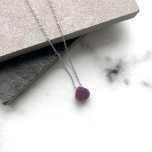 May include: A silver necklace with a small, teardrop-shaped pendant in a deep magenta hue. The pendant hangs from a fine silver chain. The necklace is displayed on a light grey surface, creating a simple and elegant look.