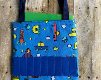 Può includere: Borsa tote blu con un motivo a tema spaziale con razzi, pianeti e stelle. La borsa ha una tasca blu e manici blu navy. Un libro verde è visibile all'interno della borsa. La borsa è in tessuto.