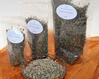 May include: Three clear plastic bags filled with dried lavender buds. The bags are labeled "Dancing Meadows Dried Lavender Buds" and are arranged on a wooden surface. A glass bowl filled with dried lavender buds is in the foreground.