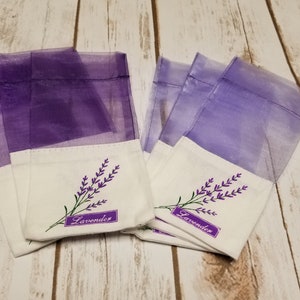 May include: Several sachets with a white base and sheer purple tops. Each sachet has an embroidered lavender sprig with the word "Lavender" on a purple label. The sachets are arranged on a light wood surface.