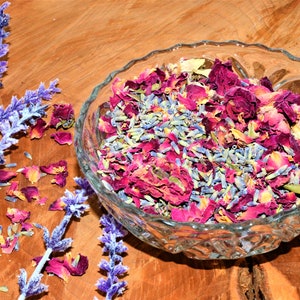 May include: A glass bowl filled with dried lavender and rose petals. The petals are a mix of pink, red, and white. The bowl is sitting on a wooden surface.