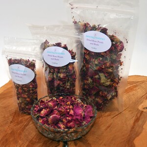 May include: Three clear plastic bags filled with dried rose petals and buds. The bags are labeled "Dancing Meadows Dried Rose Petals and Buds" with the size of the bag in ounces. A small glass bowl filled with dried rose petals and buds is in the foreground.