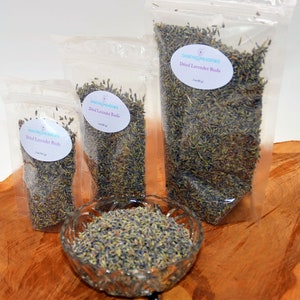 May include: Three clear plastic bags filled with dried lavender buds. The bags are labeled "Dancing Meadows Dried Lavender Buds" and have different sizes. A glass bowl filled with dried lavender buds is in the foreground.