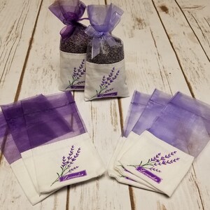 May include: Lavender sachets in organza bags with embroidered lavender sprigs. The sachets are filled with dried lavender buds and tied with purple ribbon. The bags are on a white wooden surface.