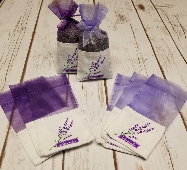 May include: Lavender sachets in organza bags with embroidered lavender sprigs. The sachets are filled with dried lavender buds and tied with purple ribbon. The bags are on a white wooden surface.