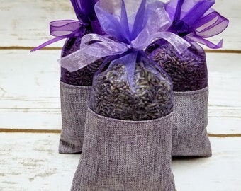 May include: Three purple fabric sachets filled with dried lavender flowers. The sachets are tied with purple ribbon and have a sheer purple overlay.
