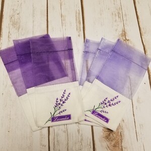 May include: Several small, sheer purple sachets with white bottoms are arranged on a light wood surface. Each sachet has an embroidered lavender sprig and the word "Lavender" on a purple label.