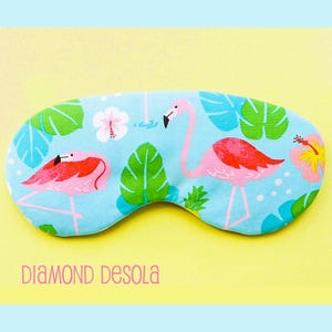 May include: A blue sleep mask with a tropical print featuring pink flamingos and green palm leaves. The mask has white polka dots and a white hibiscus flower.