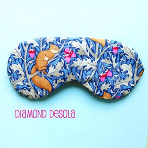 May include: A blue and beige sleep mask with a repeating pattern of squirrels, leaves, and acorns. The mask is made of fabric and has a soft, comfortable feel.