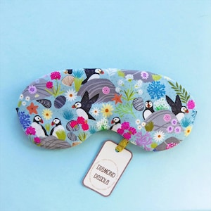 May include: A light blue sleep mask with a colorful pattern of puffins, flowers, and seashells. The mask has a tag that reads "DIAMOND DESOLA". The sleep mask is designed to block out light for better sleep.