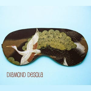 May include: Black sleep mask with a white crane flying over a gold and green pine tree design. The background is black with gold glitter.