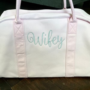 May include: A pink canvas duffel bag with two pink straps and the word "wifey" embroidered in light blue on the front.