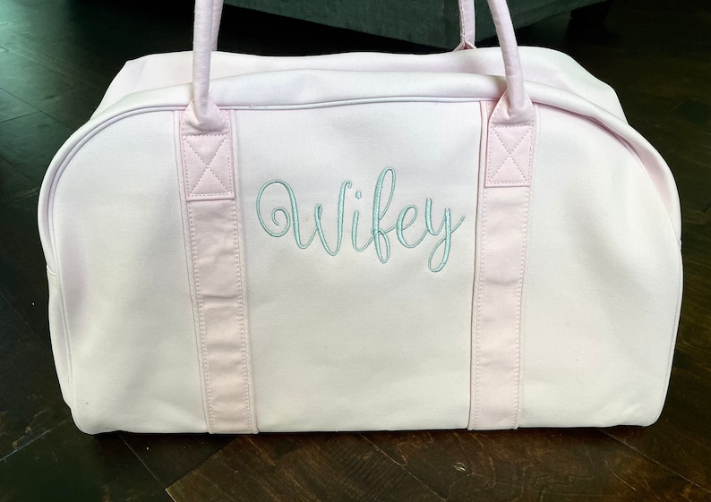 May include: A pink canvas duffel bag with two pink straps and the word "wifey" embroidered in light blue on the front.
