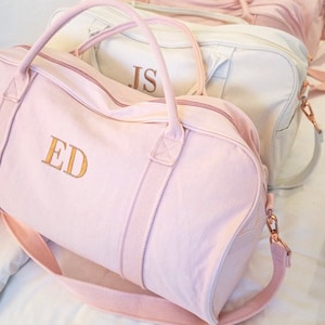 May include: Two pink canvas duffel bags with gold zipper pulls and straps. The bag on the left has the initials "ED" embroidered on the front in gold thread. The bag on the right has the initials "JS" embroidered on the front in gold thread.