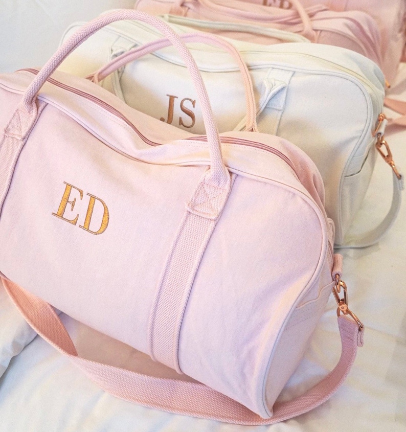 May include: Two pink canvas duffel bags with gold zipper pulls and straps. The bag on the left has the initials "ED" embroidered on the front in gold thread. The bag on the right has the initials "JS" embroidered on the front in gold thread.