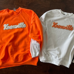 Puede incluir: Dos sudaderas con la palabra "Knoxville" en una fuente retro en la parte delantera. Una sudadera es naranja y la otra es blanca.