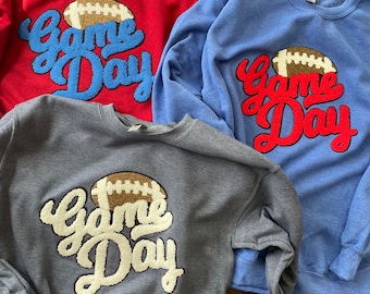May include: Three sweatshirts with the text "Game Day" in a stylized font with a football graphic. The sweatshirts are red, blue, and gray.