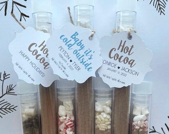 May include: Clear plastic test tubes filled with hot cocoa mix and marshmallows, each topped with a white scalloped tag. Tags feature text including Hot Cocoa, Baby it's cold outside, names, dates, and instructions to add to 1 cup of milk or water.