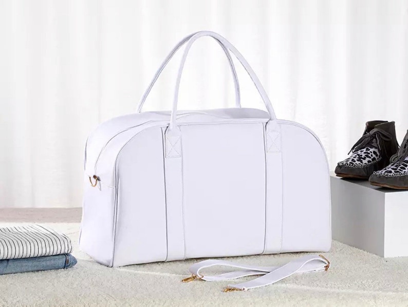 May include: White leather duffel bag with a top handle and a shoulder strap. The bag has two zipped compartments and a gold metal zipper pull.