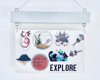 May include: A white board with a silver metal frame and string hanging from the top. The board has various stickers on it, including a black cat, a fishbowl with a fish, a cloud with rain, and a quote that says "Pull the weeds then plant the seeds". The board also has the word "EXPLORE" in large black letters.