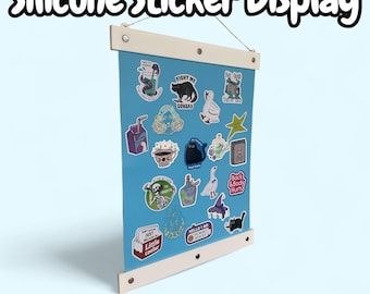 May include: A blue silicone sticker display with a white frame, showcasing various colorful stickers. The display features the text "Silicone Sticker Display" and "Reposition your stickers over and over!" Stickers include a black cat, a skeleton, and a cartoon goose.