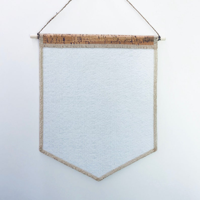 May include: A white fabric banner with a beige border and a cork rod at the top. The banner is hanging from a string.