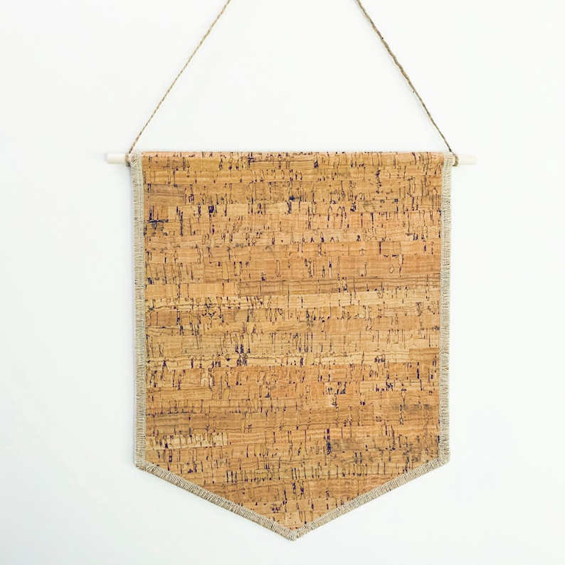 May include: A cork board banner with a white wooden dowel at the top and a light brown fabric trim. The banner is hanging from a string.