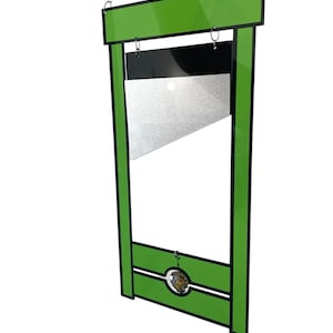 May include: A green and black metal guillotine with a silver blade. The guillotine is hanging from a chain and has a small, round, gold-colored object hanging from the bottom.